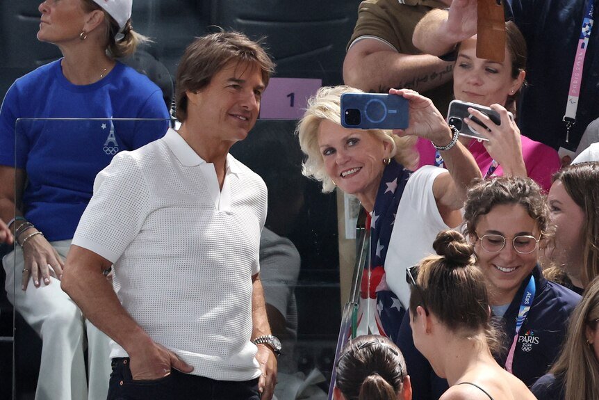 In a white polo shirt, Tom Cruise takes a selfie with a woman in the colors of the American flag